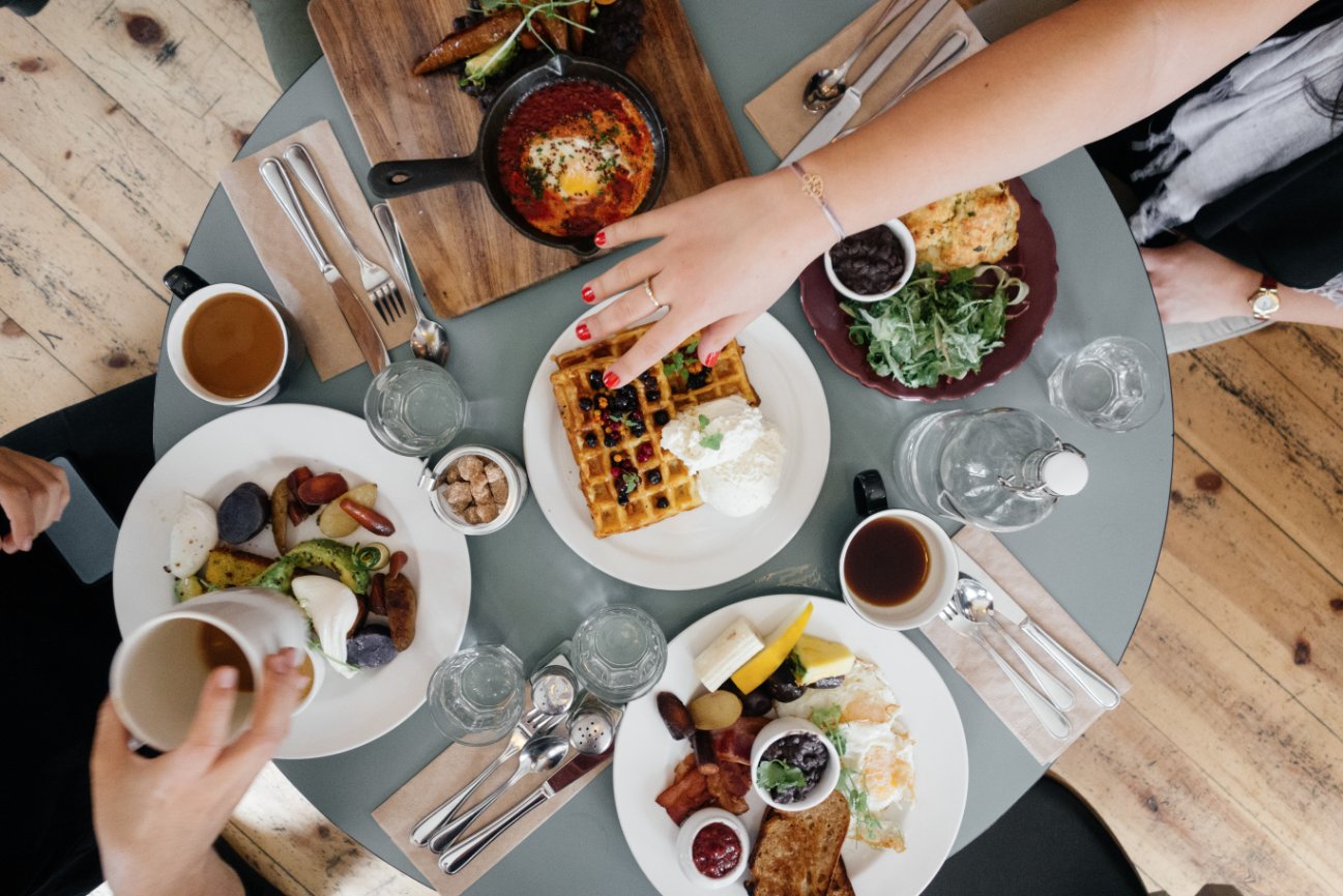 Aerial view of a dining table with waffles, eggs, bread, salad, drinks, and people reaching for food.